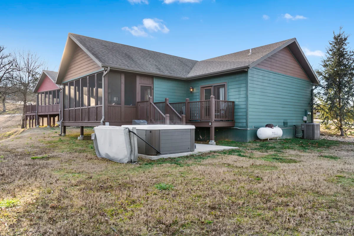 A welcoming exterior shot of the house showing its green facade, a spacious wooden deck, and a private hot tub tucked at the edge, framed by lush trees. The scene suggests a tranquil, secluded spot perfect for relaxing while taking in the surrounding nature.