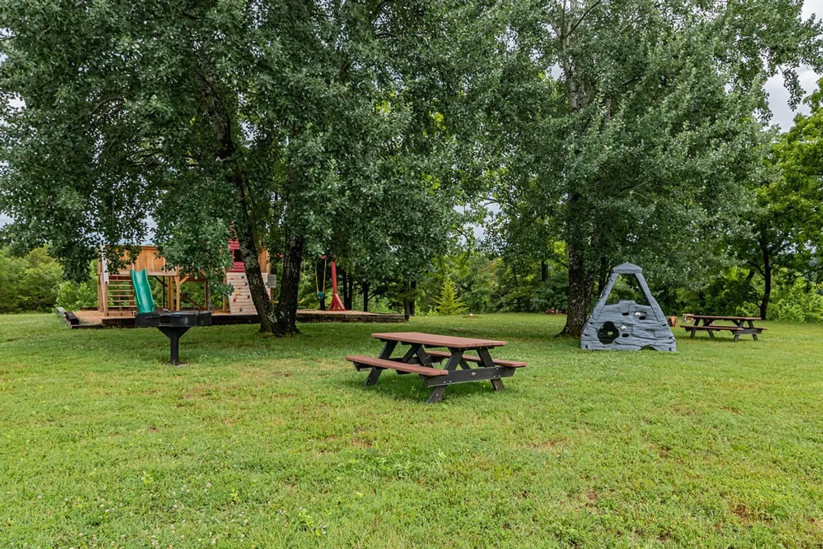 A lively exterior scene featuring a playground structure, picnic tables, a grill, and a rock climbing element, surrounded by trees. The space is designed for multi-generational enjoyment, from children playing to families barbecuing and socializing in the open air.