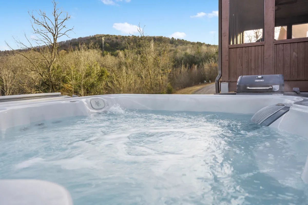 An exterior shot featuring a bubbling hot tub nestled on a patio, surrounded by lush trees and overlooking a breathtaking mountain view. The sky is vibrant with the colors of sunset, creating a serene and intimate setting ideal for relaxation and stargazing.