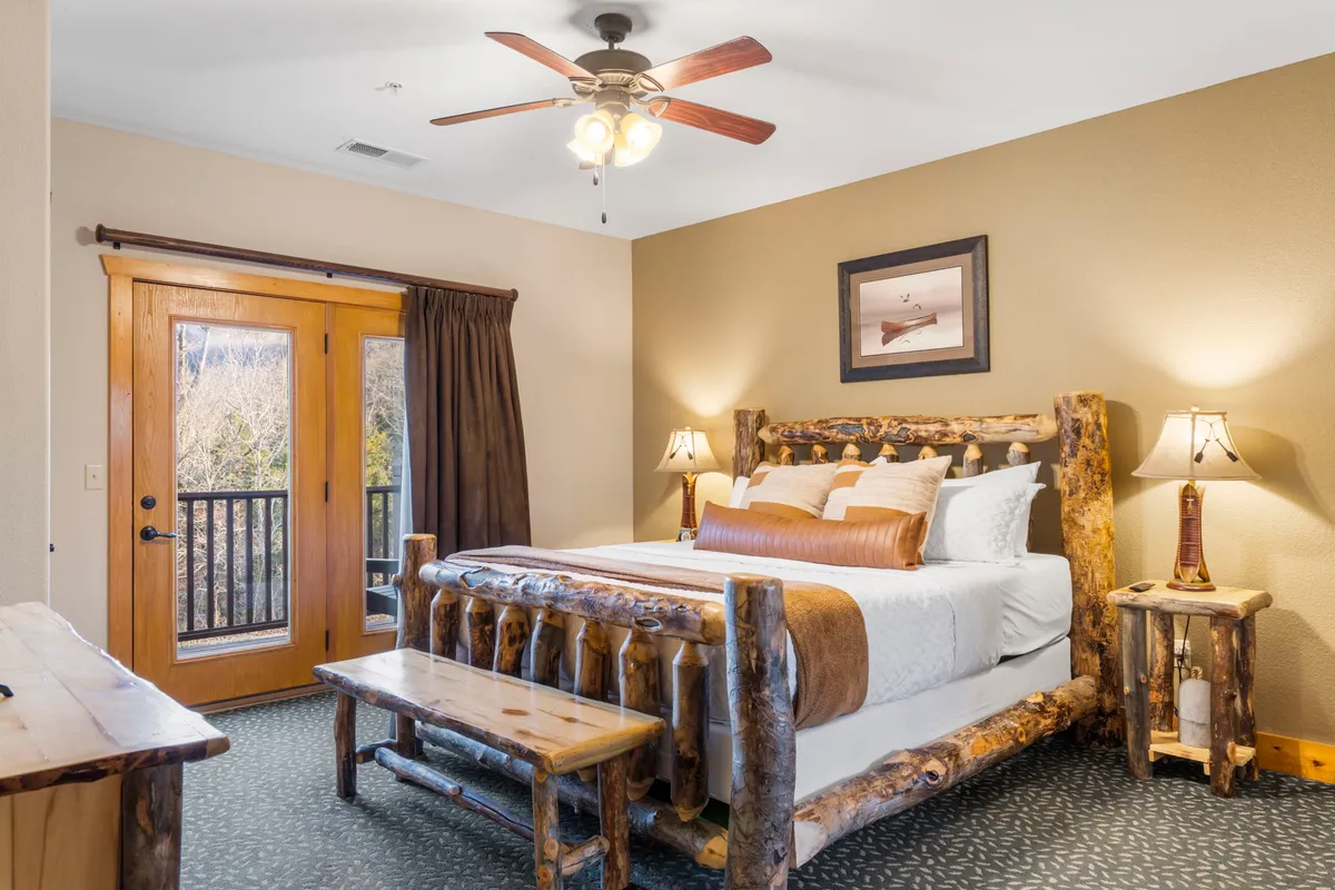 A cozy king bedroom featuring a rustic log bed, a wooden bench at the foot, decorative lamps, and a sliding door that opens onto a balcony, allowing for plenty of natural light. The ambiance is warm and welcoming, emphasizing privacy and relaxation.