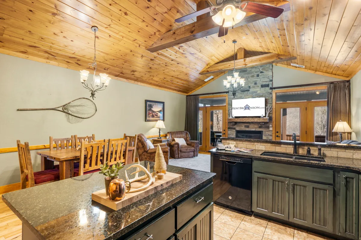 This image features an inviting interior with a central dining table, a kitchen island, a cozy seating area, and a prominent stone fireplace. The arrangement highlights the open-plan layout and communal atmosphere, where guests can gather for meals, relax, or enjoy the warmth of the fire. Large windows bring in ample sunlight, giving glimpses of the woodland surroundings.