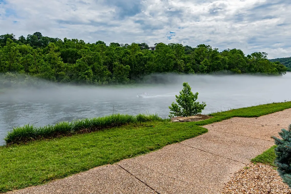 A tranquil riverside scene featuring the calm waters of the river, light morning mist drifting over the surface, lush greenery along the banks, and a winding pathway inviting visitors to stroll, fish, or simply relax in nature.