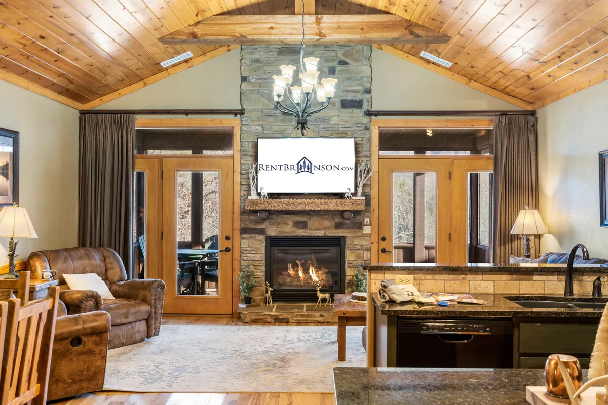 The image features a warm, inviting living room with a striking stone fireplace as the centerpiece, soft comfortable seating arranged for conversation, a rustic chandelier overhead, and large windows letting in natural light. The decor blends lodge-inspired charm with modern touches, creating a cozy and elegant space ideal for relaxing or gathering with family and friends.