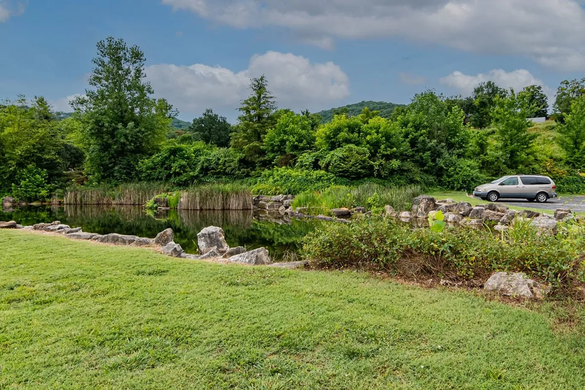 A picturesque pond bordered by vibrant greenery, natural rock formations, and distant hills, with a family-friendly atmosphere ideal for nature walks, picnics, or fishing. A parked vehicle hints at easy access for families.