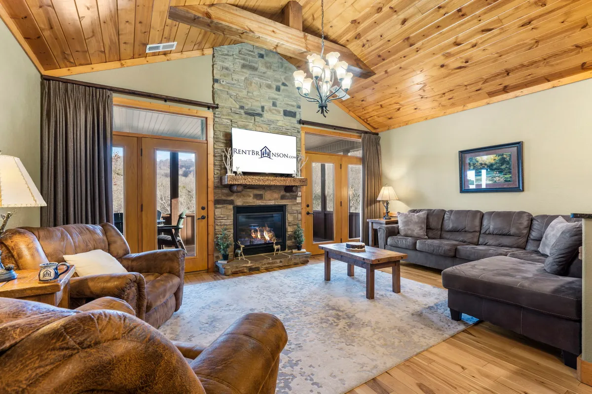This cozy living room centers on a rustic stone fireplace, with comfortable sofas and a wooden ceiling adding to the warm, welcoming atmosphere. The natural wood tones and abundant seating make it ideal for group gatherings, storytelling, or relaxing evenings indoors.