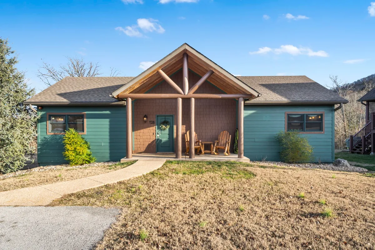This exterior image showcases the lodge's front porch with inviting rocking chairs set beneath decorative wooden beams. The green exterior walls nestle the porch comfortably into surrounding nature, offering a relaxing spot to enjoy the outdoors with friends or family.