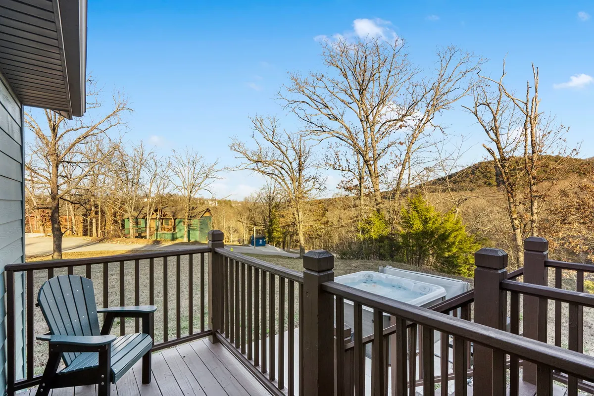 A vantage point from the wooden deck, highlighting the hot tub set amidst trees and distant hills. The deck appears spacious and peaceful, offering opportunities for stargazing, sunset watching, and immersive nature experiences.
